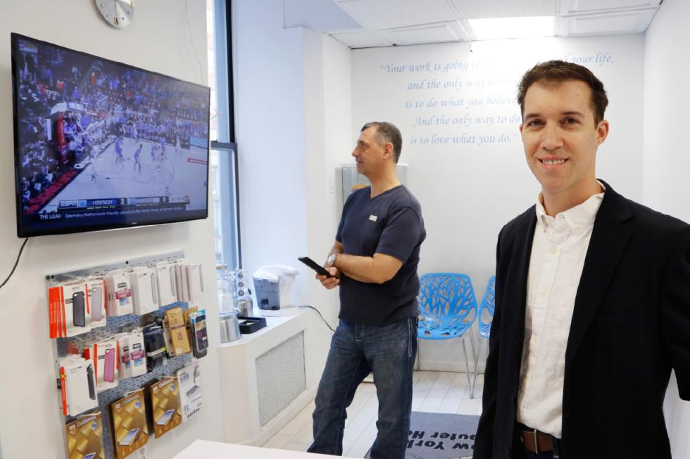 In this March 9, 2016, photo, Joe Silverman, right, poses in the lobby of his New York computer repair shop while employee Alex Lokshin watches the company's big-screen TV. Silverman realized last year he was losing productivity to the NCAA college basketball tournament, as the 20 staffers of his computer repair company kept checking their phones for scores during the games. His solution was to allow them to have the games on the big-screen TV he had installed for customers, but to let them know they do need to focus on their tasks at hand. Silverman keeps an eye on the games, too. (AP Photo/Mark Lennihan) - Mark Lennihan | AP