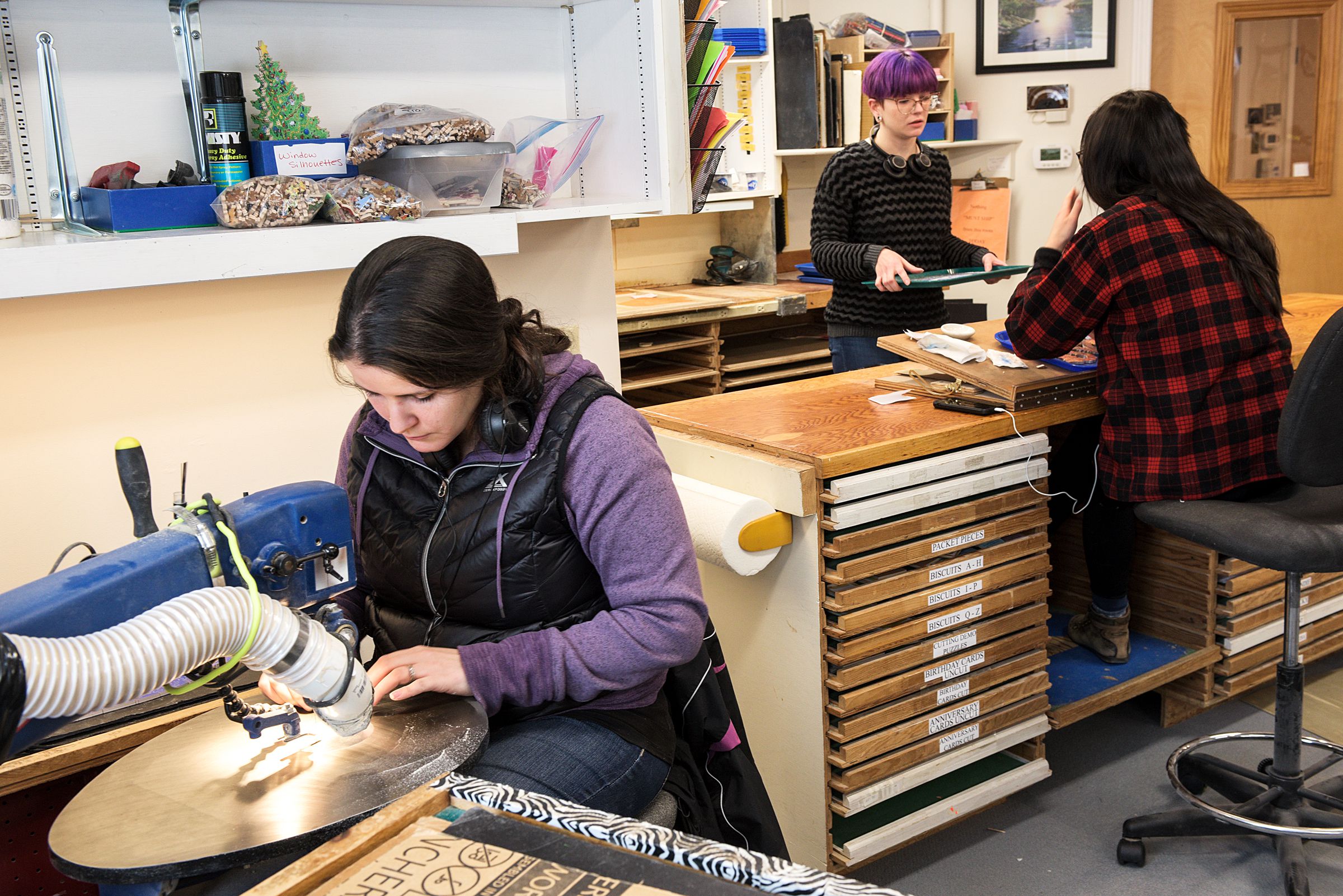 Christina Julian cuts a puzzle at left as apprentice employees Marisa Chapin, middle, and Kat Chau, right, work in the finishing area at Stave Puzzles, where pieces are touched up and the backs of the puzzles are sanded, polyurethaned and buffed in Wilder, Vt., Tuesday, Jan. 15, 2019. Apprentices work for six months to a year to learn the craft before their work can be sold. (Valley News - James M. Patterson) Copyright Valley News. May not be reprinted or used online without permission. Send requests to permission@vnews.com.