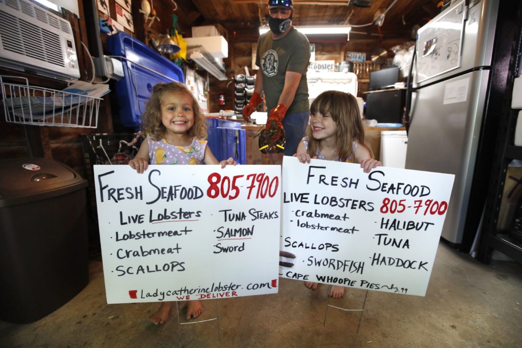 In this Friday, May 29, 2020 photo, Cora Pray, left, and her sister, Pearl Pray, hold homemade signs advertising their father's new business of selling his lobsters from his home garage in Portland, Maine. Eric Pray, background, is one of many fishermen and farmers who have pivoted quickly to sell to directly to consumers after the coronavirus shutdown cut out usual sales options. (AP Photo/Robert F. Bukaty)