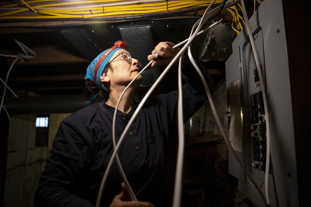 Joanna Sharf, of Cornish, N.H., guides a wire up from the basement to the first floor of a client’s house in Wilder, Vt., on Thursday, March 24, 2022. Sharf specializes in re-wiring old homes at her business Emily Electric, a name that alludes to her love of literature and poet Emily Dickinson. (Valley News / Report For America - Alex Driehaus) Copyright Valley News. May not be reprinted or used online without permission. Send requests to permission@vnews.com.