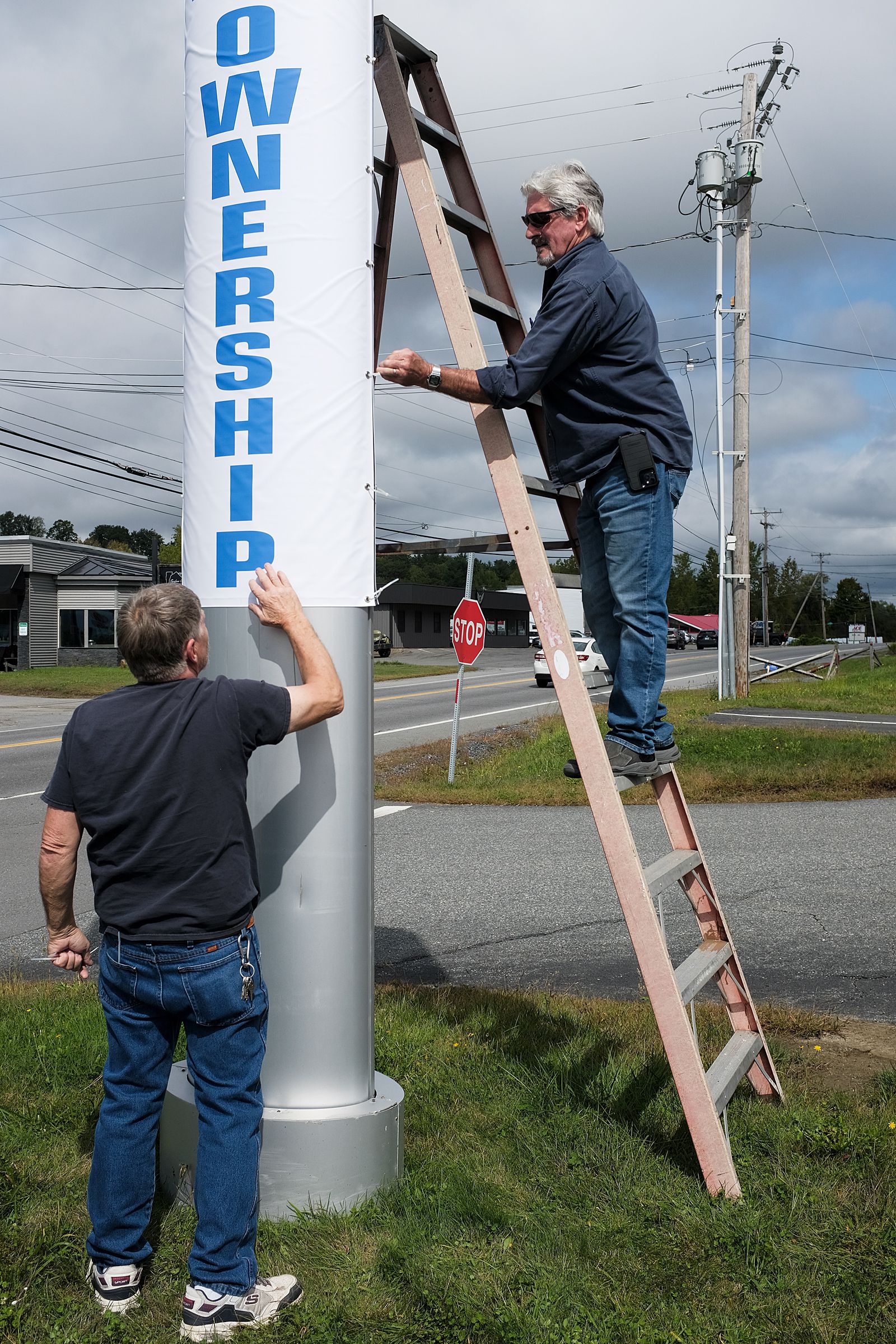 Ray St. Sauveur, of Unique Signs, right, hangs a banner with help from friend Gary Pellerin, left, at a Claremont, N.H., car dealership on Tuesday, Sept. 20, 2022. “Banners are just banners,” said St. Sauveur, who has been building, printing, welding, wiring and maintaining signs for 47 years. “Anybody can do banners.” (Valley News - James M. Patterson) Copyright Valley News. May not be reprinted or used online without permission. Send requests to permission@vnews.com.