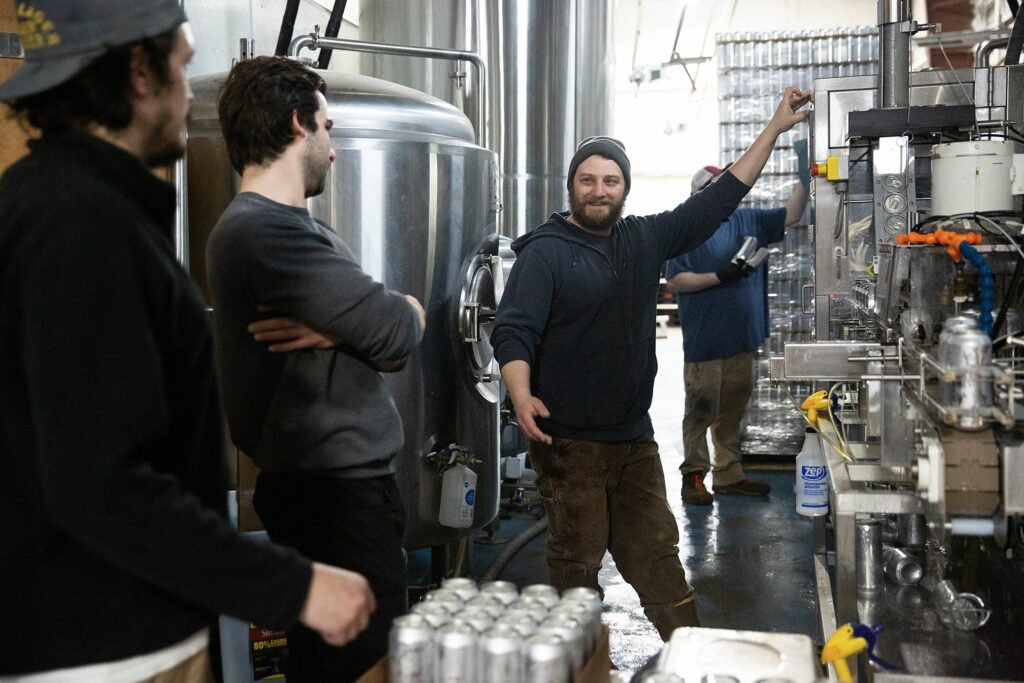 Owner Mark Babson, center, talks to Sam Clemens, left, and Kellen Van Nostrand, second from left, while Edwin Royce adds empty cans to a canning line at River Roost Brewery in White River Junction, Vt., on Tuesday, March 28, 2023. Babson started the business in 2016 and said he never imagined the small brewery would grow to its current size. (Valley News / Report For America - Alex Driehaus) Copyright Valley News. May not be reprinted or used online without permission. Send requests to permission@vnews.com.